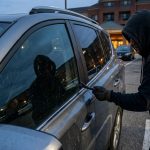Hooded thief breaking into a parked SUV in a hotel parking lot at dusk